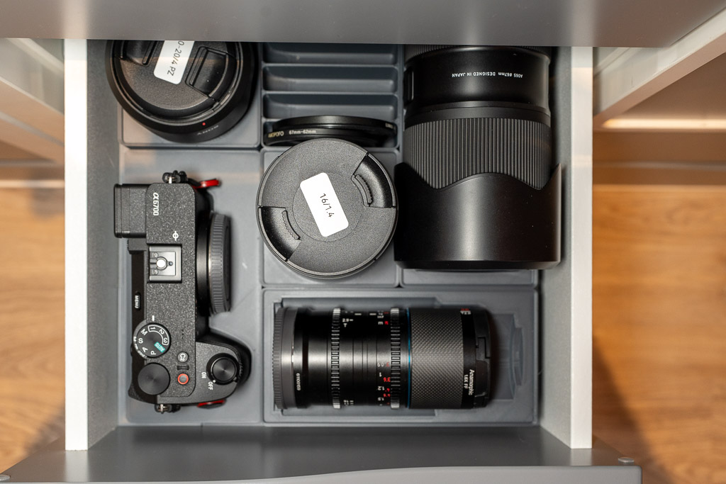 Draufsicht auf eine geöffnete graue Schublade mit perfekt eingepassten Einsätzen für Kameraequipment. Zu sehen sind eine Sony Alpha 6700, mehrere Objektive (darunter ein anamorphes Objektiv und Zoom-Linsen) sowie Filter, die sicher in grauen 3D-gedruckten Halterungen verstaut sind.