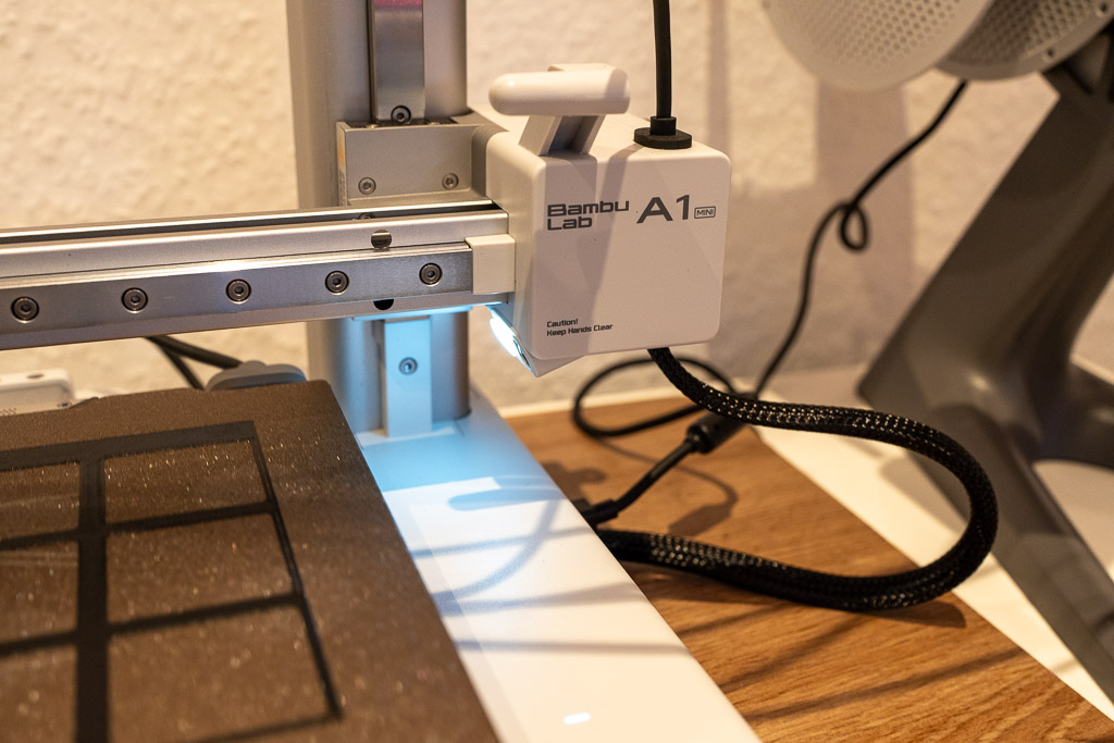 Nahaufnahme eines Bambu Lab A1 mini 3D-Druckers. Der Druckkopf mit LED-Beleuchtung befindet sich über dem Druckbett, auf dem ein schwarzes Gittermuster gedruckt wird. Im Hintergrund ist eine strukturierte weiße Wand und ein Teil des Filamenthalters zu sehen.